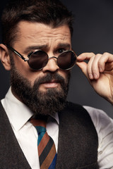 Obraz premium Low key portrait of serious handsome man with dark hair, mustache and beard in white shirt, brown vest and colorful tie takes off round sunglasses
