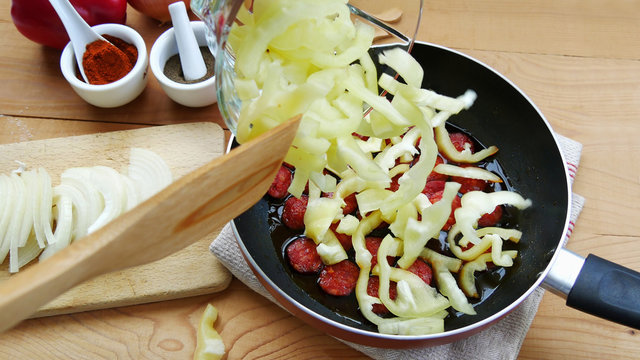 Cooking lecho with sausage, adding sliced bell peppers into the frying pan on wooden table