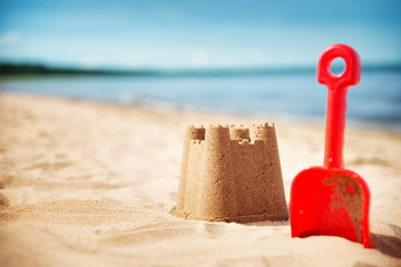 Sandcastle with a shovel on the sea in summertime. Seashore on beautiful day. Sand on the beach and blue water - Powered by Adobe