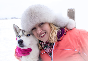 Young woman in winter clothes, smiling broadly and hugging a dog with blue eyes Husky