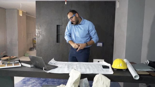 Angry man with blueprints and measuring tape talking on cellphone at new home

