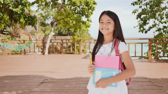 A charming filipino schoolgirl with a backpack and books in a park off the coast. A girl joyfully poses, raising her hands up with textbooks in her hands. Warm sunny day.