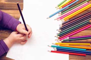 The child's hands are painted with colored pencils on a white sheet of paper on a wooden table