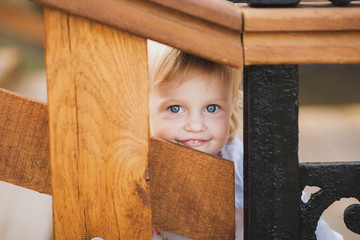 Closeup portrait of happy smiling one year old little girl looking out through wooden elements of stairs outdoors in city park. Funny cute face of beautiful small child playing on suumer day.