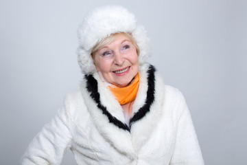 Adult smiling woman is 70 years old. An old woman in a light fur hat and fur coat against a light background.