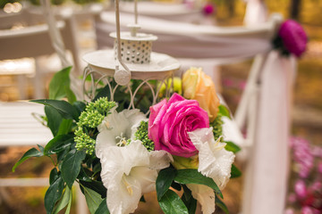 Floral elegant elements of wedding decorations. Settings for romantic wedding ceremony outside in sunny forest. White wooden empty chairs decorated with hanging lanterns full of colorful flowers.