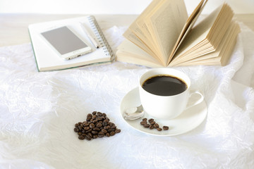 A cup of coffee, coffee beans laid out in the shape of a heart, behind an open book, notebook, mobile phone, pen on a white background.