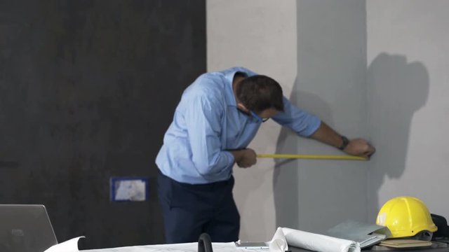 Young man with blueprints measuring wall and making notes at his new apartment 
