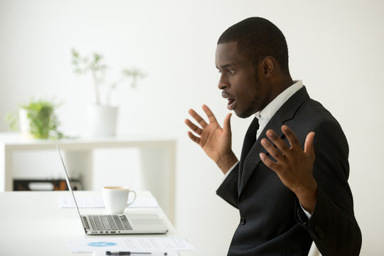 Shocked african-american businessman in suit feeling stunned by online news looking at computer screen sitting at workplace with laptop, stressed trader investor surprised by stock market changes