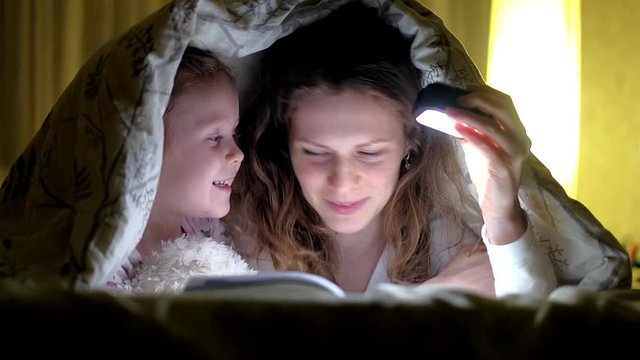 Smiling mother and her daughter reading book under bed cover and holding a flashlight.
