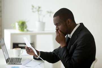 Shocked african-american businessman frustrated by notification about loan debt, confused stunned black employee feeling stressed reading unexpected bad news in letter, got dismissed fired in notice