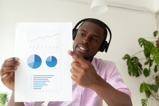 African american businessman in headphones talking by business corporate video call showing infographic statistics, black man holding document with graphs reporting distantly online, web camera view