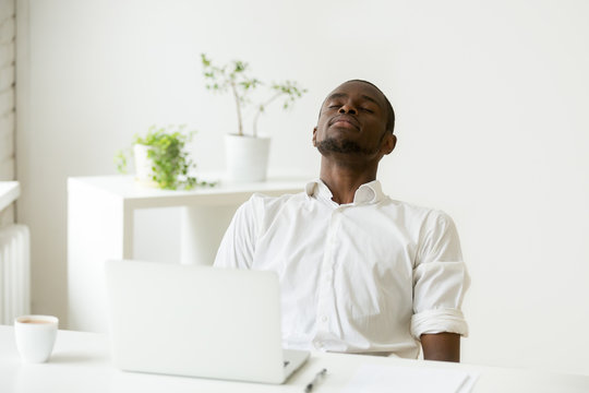 African office worker relaxing with eyes closed sitting at work desk with laptop, black employee taking rest doing exercise for relaxation at workplace during break, meditating or breathing fresh air