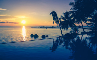 Gorgeous sunset with silhouettes of palm trees reflecting on infinite pool and calm sea on the...