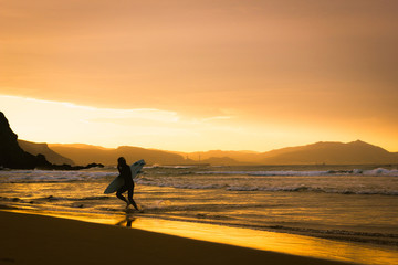 Fototapeta na wymiar Surfer with board under arm running out of water at golden hour in Atxabiribil beach, north Spain. Man enjoying quality time outdoors