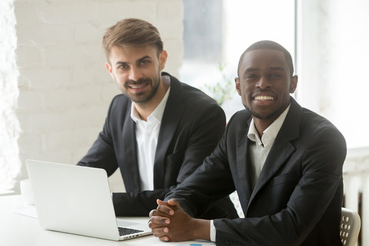 Multiracial business professionals team portrait, smiling african and caucasian businessmen in suits sitting at office desk with laptop, black and white successful startup founders looking at camera