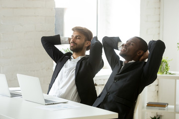 Two diverse businessmen relaxing after work breathing fresh air, relaxed african and caucasian partners in suits resting at workplace, executive corporate team enjoying break meditating in office