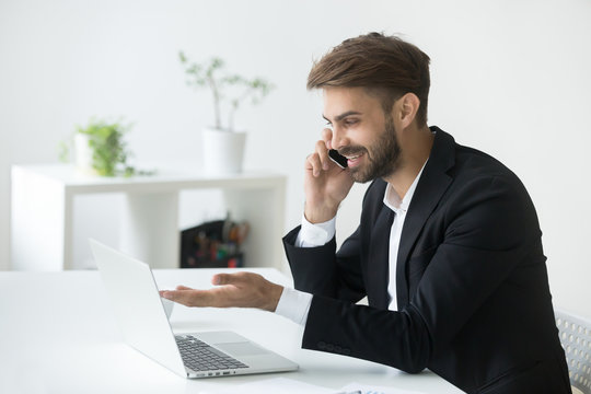 Smiling successful businessman in suit talking on phone using laptop sitting at office workplace, young entrepreneur making answering call consulting client speaking by cell, holding mobile interview