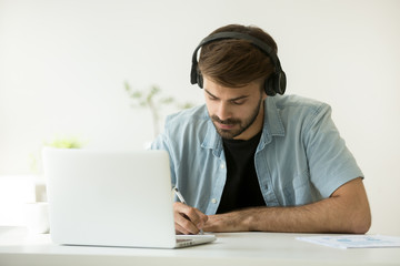 Focused man wearing headphones writing notes studying with laptop, serious student or office worker listening to audio business course at work, e-learning and online professional education concept