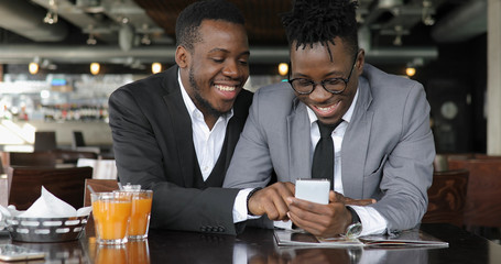 African man friends laughing in cafe, looking photos together on smart phone