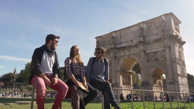 Three young friends tourists sitting in front of colosseum in rome talking having fun near arch of constantine with backpacks sunglasses happy beautiful girl long hair