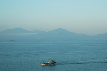 A Vietnamese boat in a bay surrounded by mountains. The flags on the boat are visible. The water, mountains and sky are all shades of blue.