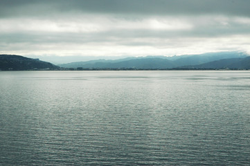 A calm sea, with mountains in the background. Storm clouds fill the sky. The water, land and sky are all shades of silver-blue.