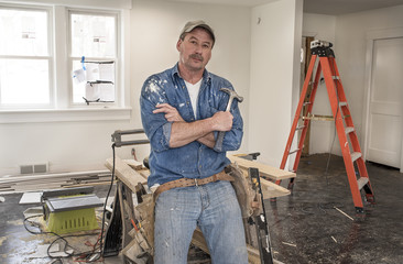Male carpenter wearing leather tool belt and holding hammer leaning against saw horse work table with chop saw in home remodel