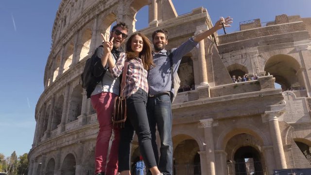 Three young friends tourists standing on pedestal in front of colosseum in rome taking funny hilarious pictures posing with backpacks sunglasses happy beautiful girl long hair slow motion