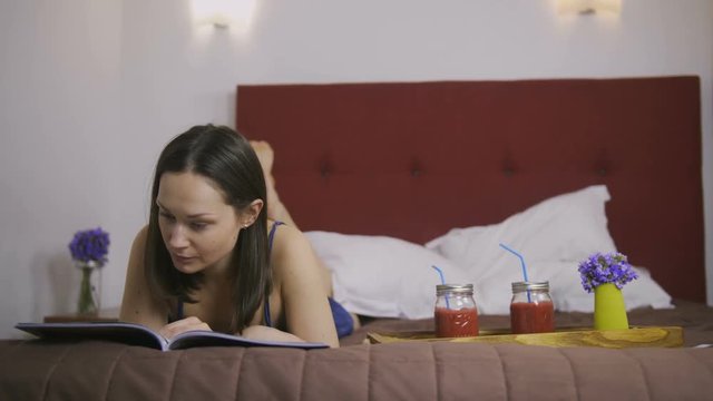 Serious young attractive brunette woman lying in bed and reading fashion magazine. Cute female reading about new fashion trends and choosing outfits. Healthy berry smoothie on wooden tray near. Dolly