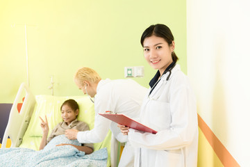 Obraz premium Happy Young asian female doctor standing and holding clipboard,background is caucasian doctor man taking care and examining patient child lying in bed in hospital. Medical and Health care kid concept.