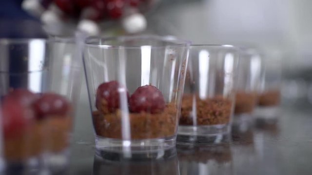 Close up of cherries being put into glasses with chokolate dough. Containers are standing in a row on black table in confectionery shop and cook is fulfilling them by the hand in white glove to make