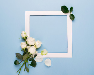  White roses and a white paper frame are decorated with fresh leaves on a blue background.Flat layout. Copy space.Celebratory concept.