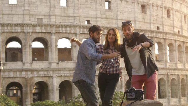 Three young friends tourists jumping in front of colosseum in rome at sunset taking selfie pictures photos with dslr camera timer happy beautiful girl long hair slow motion