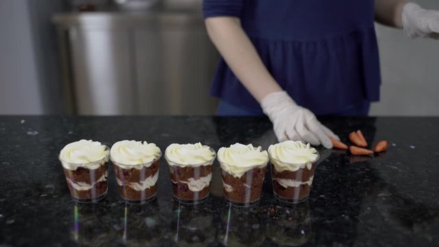 Female confectioner is decorating dessert with strawberry pieces. Five glasses with chocolate sweet bought and white cream are standing on black table in a row and woman in white gloves is putting