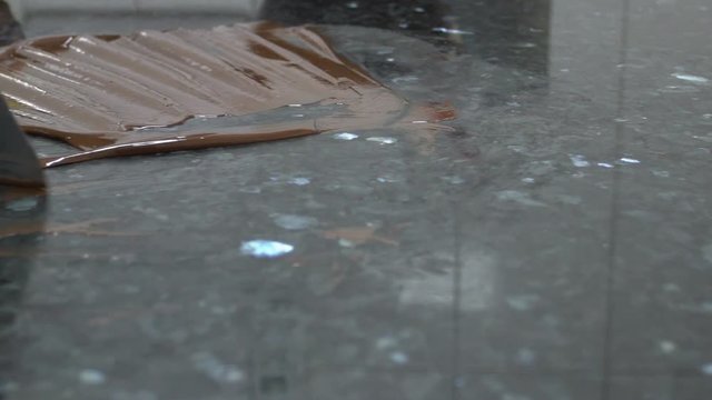 Chocolate is being deleted from the table surface on the kitchen of confectionery shop, close up. Female baker with beautiful manicure is using metal spatula to clean the grey desk from the sweet