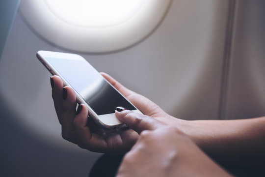 Mockup image of woman's hands holding and pointing at a white smart phone with blank black desktop screen next to an airplane window