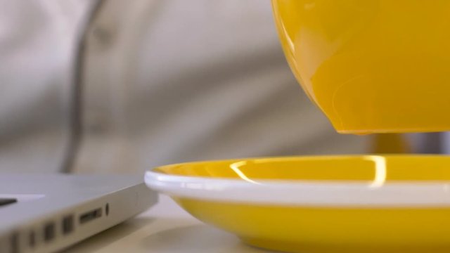 Close up of tea mug is being put onto the plate in moderm cafe. Cup with hot tasty beverage is yellow and standing on wooden table near brand-new laptop, on which client in light shirt is working