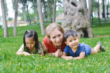 Fototapeta premium Happy biracial family in the summer park - caucasian mother with her latin children laying on the grass.