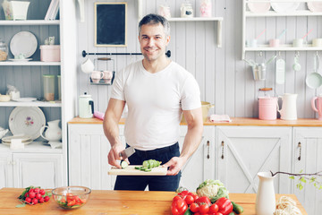 Portrait of handsome smiling man at kitchen. cooking and home concept - close up of male hand chopping cucumber on cutting board with sharp knife