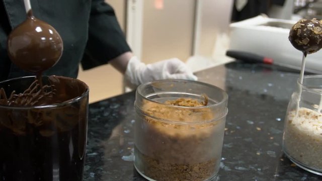 Tasty lollypop is being cooked on the kitchen of confectionery shop. Professional in black uniform and white gloves is putting the candy to the melted chocolate, than applying flackes from glass can
