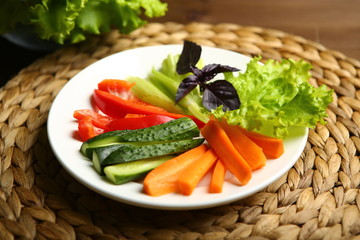 Pepper, carrots, celery, raw cucumber are cut into strips. On the plate. Next to it are lettuce leaves, basil. On a background a wicker stand, a wooden table, a black background.