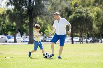 Tuinposter young happy father and excited little 7 or 8 years old son playing together soccer football on city park garden running on grass kicking the ball © Wordley Calvo Stock