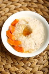Low-calorie porridge for a healthy diet and lifestyle. Barley with honey and bits of orange pumpkin in a deep white plate. Dark background, wooden table and wicker stand