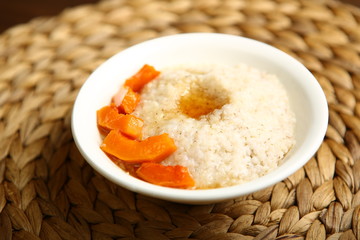 Low-calorie porridge for a healthy diet and lifestyle. Barley with honey and bits of orange pumpkin in a deep white plate. Dark background, wooden table and wicker stand