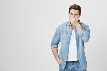 Studio shot of trendy young guy with stylish haircut laughing and trying to hide it, covering mouth with hand, standing over gray background. Man feels sick after eating something