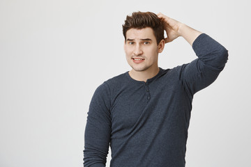 Indoor shot of cute athletic man in casual clothes, scratching back of head with question and confused expression, standing over gray background. I am sorry mom, I forgot take chicken out of fridge