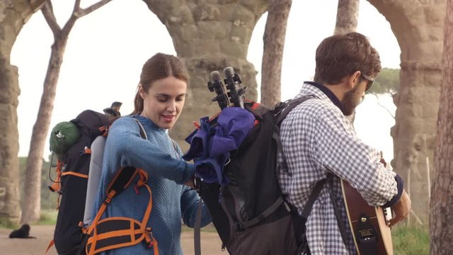 Young couple backpackers tourists searching fixing open backpack near roman aqueduct arches in parco degli acquedotti park ruins in rome at sunrise with guitar and sleeping bag slow motion steadycam