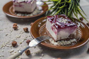 A piece of vanilla cake with chocolate base and cherry glaze decorated with some pieces of white chocolate served in a brown clay dish with silver fork placed on a beige tablecloth. Blurred background