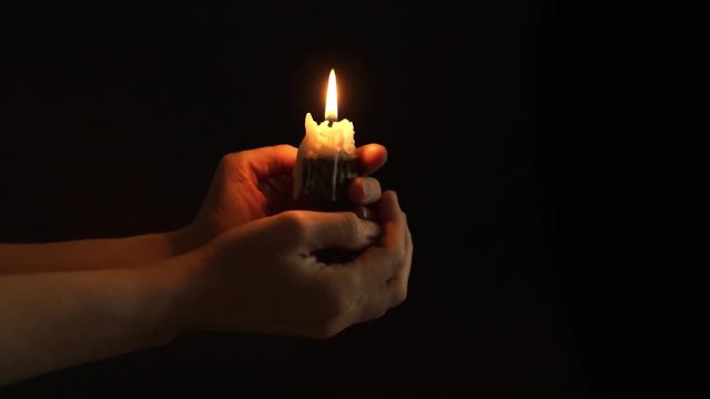 Single lit white candle in candlestick with waxy stains close up a dark background. Hands holding candle in dark black background, the flames sway from the breath, at the end it blows out and goes out